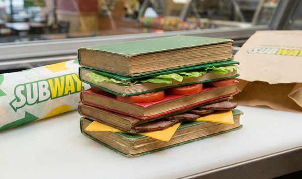 A sandwich constructed from vintage books layered with lettuce, tomatoes, meat, and cheese.